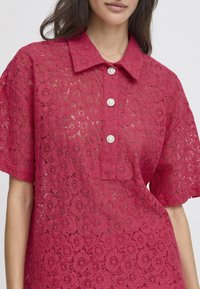 Blusa rossa in pizzo con collo classico, maniche corte e tre bottoni simili a perle. Presenta un motivo floreale e una trama trasparente.