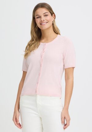 Donna sorridente con capelli lunghi che indossa un cardigan rosa chiaro a maniche corte con bottoni e pantaloni bianchi, in piedi davanti a uno sfondo semplice.
