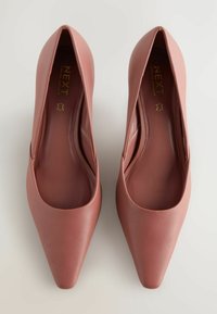 Tacchi in pelle rosa a punta, con una texture liscia, caratterizzati da un design semplice e dettagli minimalisti, etichettati "NEXT Forever Comfort."