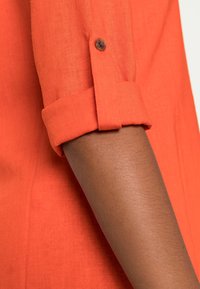 Camicia di lino arancione con maniche arrotolate, caratterizzata da un bottone marrone visibile su un tessuto testurizzato. Primo piano di un braccio che indossa la camicia.
