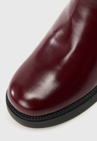 Bota de tornozelo de couro bordeaux com um acabamento brilhante, bico arredondado e sola de borracha preta com detalhes texturizados ao longo da borda.