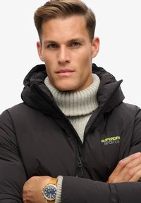 Schwarze Pufferjacke mit Kapuze und gelbem Logo, kombiniert mit einem hellgrauen, gerippten Rollkragenpullover. Trägt eine Uhr mit braunem Armband.