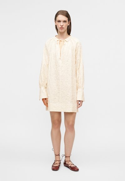 Φόρεμα μήκους μέχρι το γόνατο σε ανοιχτό μπεζ χρώμα με υφή, γιακά και δέσιμο στη λαιμόκοψη. Διαθέτει μακριά μανίκια και γραμμή Α.