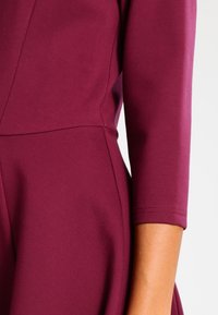 Läheltä katsottuna henkilö, joka on pukeutunut sääntöjen mukaan istuvaan, pitkähihaiseen violettiin mekkoon, jossa näkyy käsivarren ja vyötärön yksityiskohtia sekä saumoja.