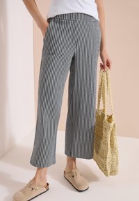 Personne portant un pantalon large à rayures verticales bleues et blanches, des chaussures beiges sans lacets, tenant un sac cabas beige au crochet.