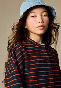 Gestreepte trui in marineblauw en oranje, gemaakt van zachte stof met een kraag met knopen. Draagt een blauwe bucket hoed.