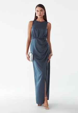 Vrouw in een mouwloze, tot op de vloer reikende staalblauwe jurk met zijsplit, zilveren clutch, zilveren sandalen met hoge hakken en bijpassende oorbellen en armband.