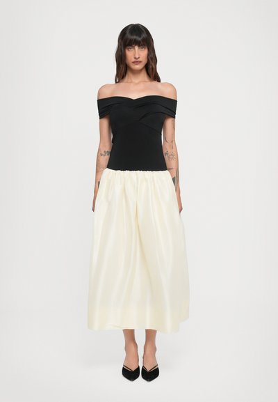 Off-the-shoulder sort top, plisseret elfenbenskørt, indsnævret talje, glat tekstur. Sorte spidse hæle. Enkel og moderne design.