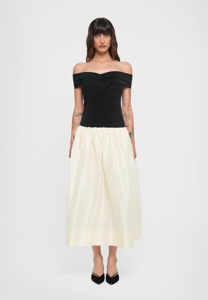 Off-the-shoulder sort top, plisseret elfenbenskørt, indsnævret talje, glat tekstur. Sorte spidse hæle. Enkel og moderne design.