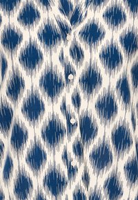 Biela košeľa s modrým nepravidelným kruhovým vzorom štetcom a prednými gombíkmi, detail na oblasť trupu.