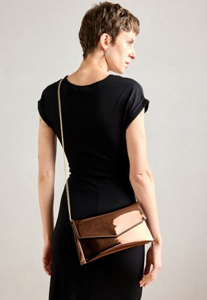 Woman with short hair wearing a black dress and metallic bronze crossbody purse with gold chain strap, facing sideways against a plain background.