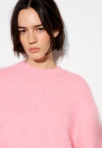 Tânără cu păr scurt și închis la culoare, purtând un pulover moale roz pal, pozând pe un fundal alb simplu.