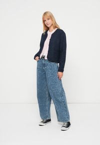 Marineblå cardigan med tekstur over en lys rosa knappet bluse, kombinert med lyseblå vidbeinte jeans med slangeskinnsmønster og svarte joggesko.