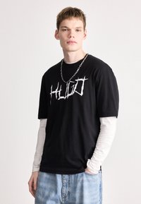 Μαύρο βαμβακερό T-shirt με λευκό γραφικό λογότυπο "HUGO", που φοριέται πάνω από ένα λευκό μακρυμάνικο πουκάμισο, συνδυασμένο με ανοιχτό μπλε τζιν παντελόνι.