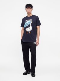 Νεαρός άνδρας με μαύρο μπλουζάκι Moschino με λευκή πάπια που κρατάει ένα μπλε ομπρέλα, μαύρο παντελόνι Moschino και μαύρα παπούτσια, στέκεται με τα χέρια στις τσέπες.