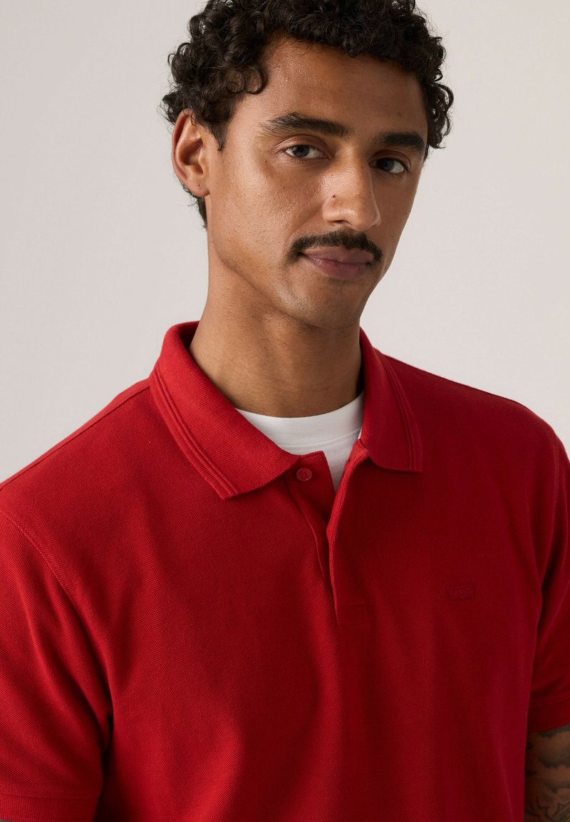 Homme aux cheveux bouclés et moustache, portant un polo rouge par-dessus un t-shirt blanc, regardant légèrement sur le côté sur un fond neutre.