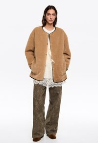 Mujer vestida con una chaqueta de forro polar crema abotonada, blusa de encaje blanca, pantalones de pierna ancha con estampado, zapatos marrones, con las manos en los bolsillos de la chaqueta, de pie recta.