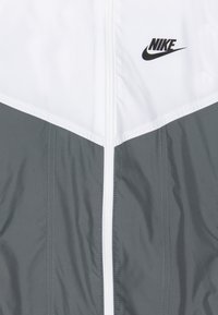 Nike zipová bunda v šedé a bílé barvě, s designem šipky, hladkou texturou a černým logem na prsou.