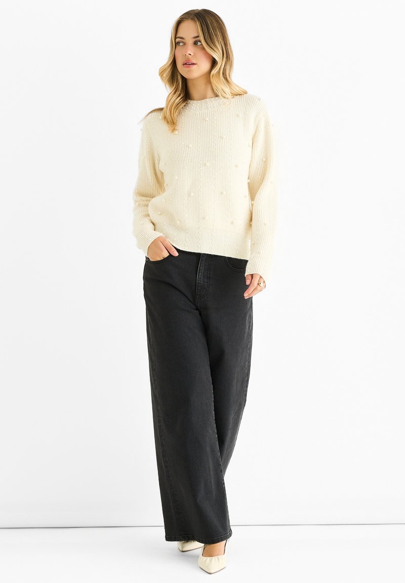 Cream-colored knit sweater with textured bobble details, paired with loose black denim pants and light-colored pointed-toe shoes.