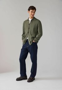 Cardigan tricotat de culoare verde măslin, cu cusături decorative, închidere cu nasturi și două buzunare frontale, purtat cu pantaloni din denim închis și pantofi negri.