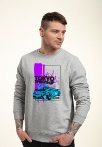 Felpa grigia con una grafica colorata di un'auto sportiva, accentuata da schizzi di vernice viola e blu e il testo "TOKYO SPEED SHOP".
