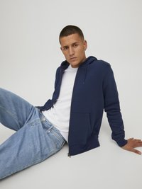 Sudadera con cremallera de color azul marino en tela suave, con un bolsillo tipo canguro, llevada sobre una camiseta blanca y combinada con unos jeans de mezclilla azul claro.