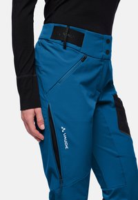 Blauwe buitensportbroek van Vaude met een aansluitend ontwerp, ritszakken aan de zijkanten, gestructureerde zwarte accenten en een logo op het linkerbeen.