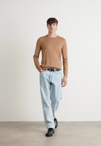 Light brown knitted sweater, blue loose-fitting jeans, black shoes. Model stands on a neutral background, hands in pockets, relaxed posture.
