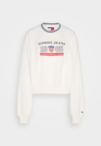 Biała krótkia bluza z klasycznym okrągłym dekoltem, z napisem "TOMMY JEANS 1985 INTERNATIONAL GAMES" w granatowym i czerwonym kolorze. Miękka tkanina, długie rękawy.