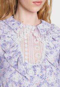 Mladá žena v levandulových květinových šatech s bílými krajkovými detaily a volánkovým límcem, ukazuje krk a částečně obličej.