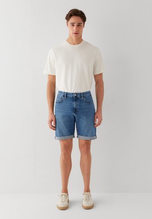 Junger Mann steht vor einer weißen Wand, trägt ein weißes T-Shirt, blaue hochgekrempelte Jeansshorts und weiße Turnschuhe, schaut nach vorne mit entspannten Armen.