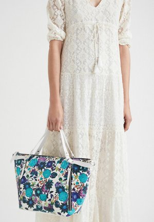 Blomsterhandtaske med turkis og lilla mønstre, hvide accenter, lynlåslukninger og dobbelte håndtag, parret med en blond, cremefarvet kjole.