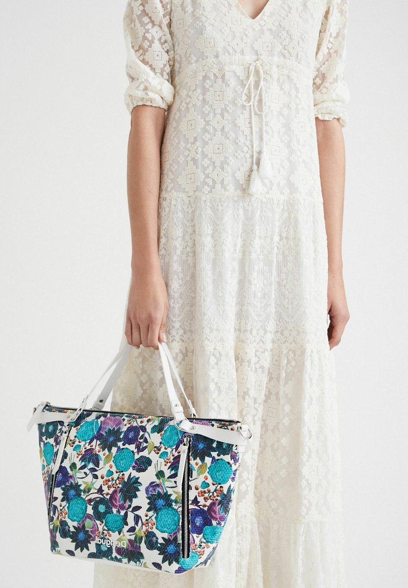 Kwiatowa torebka z turkusowymi i fioletowymi wzorami, białymi akcentami, z zamkiem błyskawicznym oraz podwójnymi uchwytami, w połączeniu z koronkową, kremową sukienką.