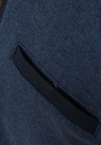 Blaues Textil mit einer dunkelblauen Einschubtasche. Hat eine glatte Oberfläche und ein dezentes Muster. Die Tasche hat ein kreisförmiges, schwarzes Knopfdetail.