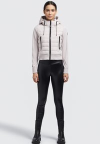 Hellgraue, taillierte Jacke mit Kapuze und Reißverschluss vorne, mit schwarzen Akzenten und zwei Reißverschlusstaschen. Getragen mit schwarzen Leggings und Stiefeln.