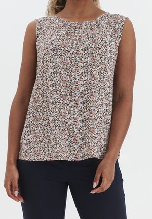 Bluzka bez rękawów w kwiaty w kolorach różowym, białym i czarnym, noszona z ciemnogranatowymi spodniami na stojącej kobiecie.