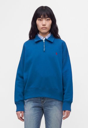 Femme aux longs cheveux noirs porte un pull bleu à demi-fermeture éclair sur une chemise bleu clair à col et un jean bleu délavé, debout sur fond blanc.