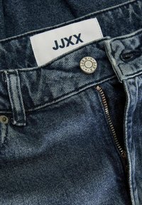 Tumši zila džinsu auduma bikses ar pogu, rāvējslīzi un baltu etiketi, uz kuras ir uzšūts "JJXX", iekļauta jostasvietā. Nedaudz nodilusi tekstūra.