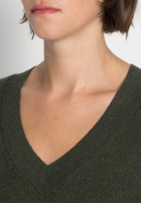 Grüner, gestrickter V-Ausschnitt-Pullover mit einem strukturierten Stoff und einem gerippten Ausschnitt. Nahaufnahme des Schulter- und Halsbereichs.