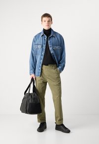 Jeans jakna z dvema žepoma na prsih, črna turtleneck, olivno zelene hlače in črne superge. Drži črno športno torbo.