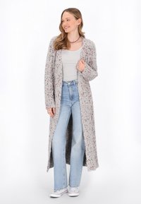 Lång, grå cardigan med strukturerad stickning, med flerfärgade fläckar. Öppen front, ribbade ärmslut och sidofickor. Bärs över en ljus skjorta och blå jeans.