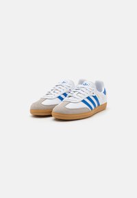 Hvite atletiske sko med blå striper og beige toecap i semsket skinn. Gummisåle, snøresystem og logo på tungen.