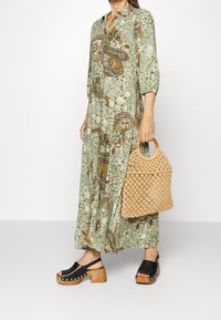 Lungo abito verde con motivo paisley, maniche a tre quarti; abbinato a zoccoli neri e a una borsa intrecciata beige con manici di legno.