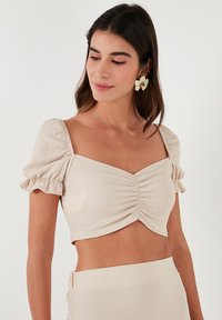 Beige īsais tops ar kvadrātveida apkakli, pūkainām piedurknēm un sakoptu priekšpusi, izgatavots no strukturēta auduma. Nēsāts kopā ar atbilstošām svārkiem.