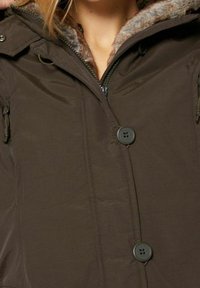 Brun isolerad jacka med huva klädd i fuskpäls, framsida med dragkedja, två sidofickor och framträdande svarta knappar.