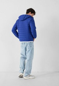 Giacca imbottita blu con cappuccio, caratterizzata da cuciture verticali, indossata sopra una camicia bianca e abbinata a jeans azzurri e sneakers grigie.
