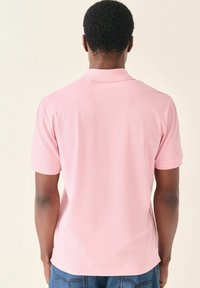 Camisa polo rosa de manga corta y con cuello, hecha de tela suave. Lleva encima de unos vaqueros de mezclilla azules, vista desde la parte de atrás.