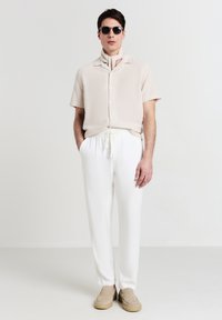 Camicia corta beige chiaro a maniche corte con colletto, abbinata a pantaloni bianchi con coulisse e scarpe beige slip-on. Occhiali da sole neri indossati.