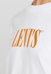 Valkoinen puuvillainen T-paita, jossa on näyttävä oranssi "LEVI'S" -logo. Klassinen pyöreä kaula-aukko, sileä pinta ja lyhyet hihat.