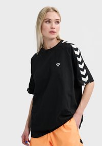 Zwarte oversized t-shirt met een ronde halslijn, voorzien van witte chevronstrepen op de mouwen en een klein logo op de borst.